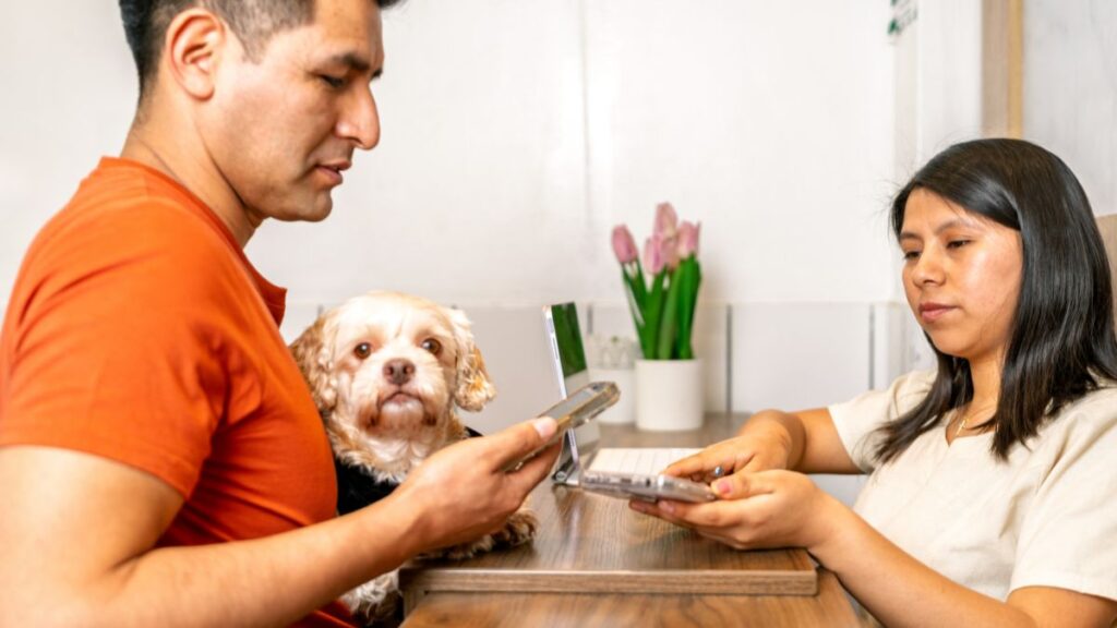 Planejamento da vacinação em pets: duas pessoas junto a um cachorro, com celular e prancheta sobre uma mesa.
